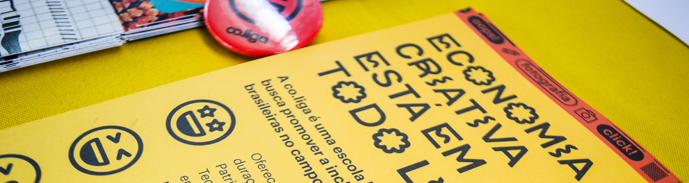 Material gráfico promocional sobre economia criativa disposto sobre uma superfície amarela. Em destaque, um folder amarelo com o título "ECONOMIA CRIATIVA ESTÁ EM TODO LUGAR" escrito com letras estilizadas. Abaixo, há um texto explicativo sobre a iniciativa Co.liga, uma escola livre digital gratuita voltada à inclusão produtiva de juventudes no campo da economia criativa. Em cima do folder, estão bottons coloridos, sendo o principal um azul com ícone de câmera e o nome "Co.liga", indicando uma abordagem vol