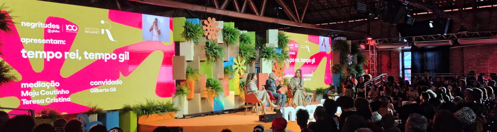 Auditório lotado assiste ao painel "Tempo Rei, Tempo Gil", durante o evento Negritudes Globo. No palco, decorado com plantas e painéis coloridos, estão sentados Gilberto Gil, a jornalista Maju Coutinho e a cantora Teresa Cristina, que conduzem a conversa. Um telão ao fundo exibe o nome do painel, os nomes dos participantes e as marcas dos organizadores. O ambiente é coberto, com estrutura de madeira e iluminação quente. 