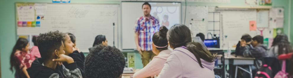 Sala de aula com diversos estudantes sentados em grupos ao redor de mesas, atentos ao professor que está em pé à frente da sala, próximo a um quadro branco com projetor. O ambiente é colorido e decorado com cartazes e materiais didáticos. Os alunos, de diferentes origens, estão concentrados na explicação, demonstrando engajamento com a atividade.