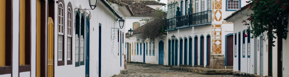 Uma rua de pedras irregulares, típica do centro histórico de Paraty, no Rio de Janeiro, aparece vazia em um dia nublado. De ambos os lados, há casarões coloniais bem preservados, com paredes brancas, portas e janelas coloridas em tons como amarelo, azul, verde e vermelho. As janelas têm grades e caixilhos de madeira, e há postes de luz com design antigo fixados nas fachadas. Um dos prédios se destaca com detalhes ornamentais em amarelo e branco e sacada de ferro. À direita, uma trepadeira com flores vermelh