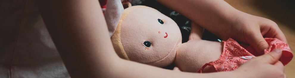 Criança branca sentada com boneca de pano no colo. O rosto da menina não é mostrado na imagem.