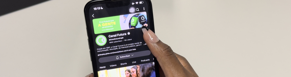 Uma mão feminina de pele negra segura um celular, que mostra a tela do canal “Canal Futura” no aplicativo do YouTube. A tela exibe o perfil do canal, com o logotipo verde circular e o texto de descrição, além de uma miniatura de vídeo com três jovens estudantes manipulando um objeto em sala de aula. A interface do YouTube está no modo escuro. O fundo é uma superfície branca, lisa e iluminada.