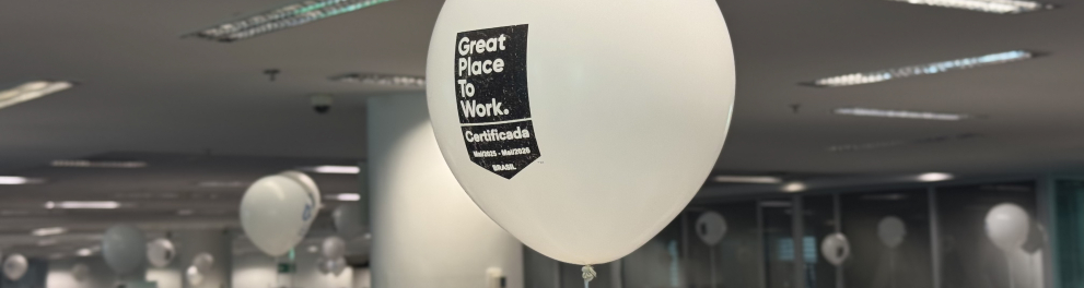 Ambiente interno de escritório com diversas pessoas concentradas em seus computadores, trabalhando em um espaço moderno, iluminado e com layout aberto. No centro da imagem, um balão branco com a frase “Great Place To Work – Certificada – Mai/2023 - Mai/2025 – BRASIL” em destaque, indicando o reconhecimento da empresa como um excelente local para trabalhar. Ao fundo, vê-se o logo estilizado da instituição em grande escala na parede e funcionários ocupando diferentes estações de trabalho. O clima é de rotina 