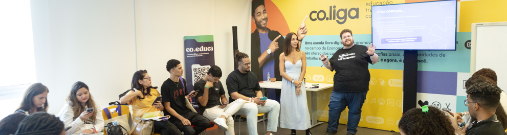 Cerca de 20 pessoas participam de uma oficina realizada em uma sala clara e colorida, com paredes decoradas com as marcas “co.liga” e “co.educa”. A maioria dos participantes está sentada em cadeiras amarelas e vermelhas, organizadas em semicírculo, com atenção voltada para dois facilitadores em pé ao centro — uma mulher de vestido claro e um homem com camiseta preta onde se lê “Transformando realidades”. Muitos usam celulares ou fazem anotações. À direita, uma tela exibe a frase “E pra começar...”, com inst