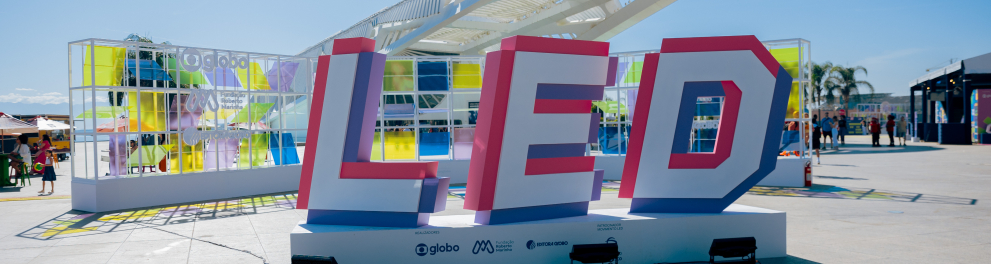 Em uma ampla área externa sob céu azul e ensolarado, destaca-se uma grande instalação com as letras tridimensionais “LED” em cores vibrantes (rosa, branco, roxo e azul), posicionada diante do Museu do Amanhã, no Rio de Janeiro. Ao fundo, uma estrutura colorida com painéis translúcidos em tons de amarelo, verde, azul e roxo exibe os logotipos da Globo, Fundação Roberto Marinho e Editora Globo, realizadores do evento.