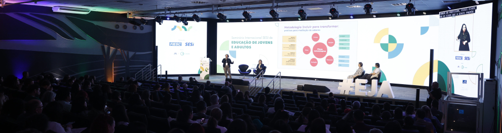 Auditório moderno com cadeiras ocupadas por diversas pessoas assistindo a uma apresentação. No palco iluminado, há quatro pessoas — duas em pé e duas sentadas — participando de um painel com telão ao fundo. No telão está escrito: “Seminário Internacional SESI de Educação de Jovens e Adultos”, com gráficos e textos explicativos. À direita, uma tela vertical mostra uma intérprete de Libras (Língua Brasileira de Sinais). O ambiente tem iluminação colorida nas laterais e teto com painéis de LED.