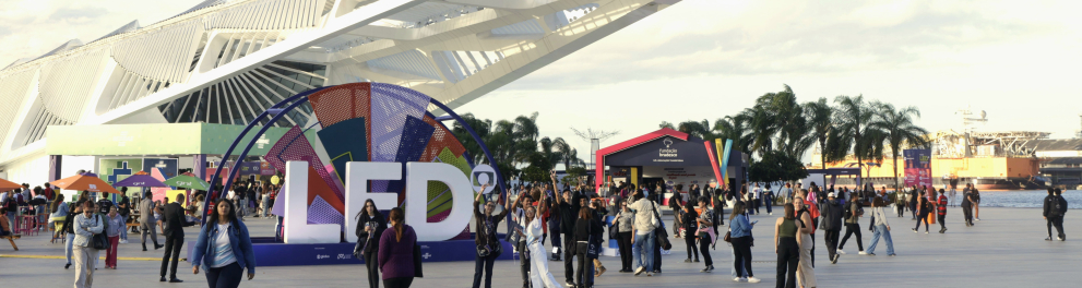 Vista ampla da área externa do Museu do Amanhã, no Rio de Janeiro, durante o Festival LED – Luz na Educação. Em primeiro plano, pessoas caminham e interagem ao redor de uma grande instalação colorida com as letras “LED”. Ao fundo, o mar, palmeiras e tendas coloridas compõem o cenário festivo.