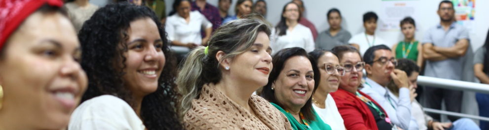 Auditório cheio com pessoas sentadas e sorrindo. Na frente, mulheres de diferentes idades assistem à apresentação. Elas demonstram atenção e simpatia. Ao fundo, mais pessoas estão em pé, observando. A sala é bem iluminada, com janelas grandes e cartazes nas paredes. O ambiente é acolhedor e participativo.