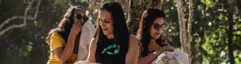 Três mulheres sentadas na areia, à sombra de árvores, em um momento de bordado coletivo. Todas estão concentradas em seus trabalhos, segurando panos com bordados coloridos. Uma usa vestido amarelo, outra usa calça estampada em preto e branco, e a terceira, um vestido floral vermelho. A luz do sol entra pelas árvores, criando uma atmosfera acolhedora e tranquila.