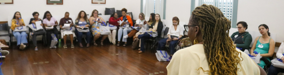 Um grupo de pessoas está sentado em círculo em uma sala. Uma mulher de pé, no centro, fala com o grupo e sorri. A sala tem piso de madeira e quadros retangulares nas paredes.