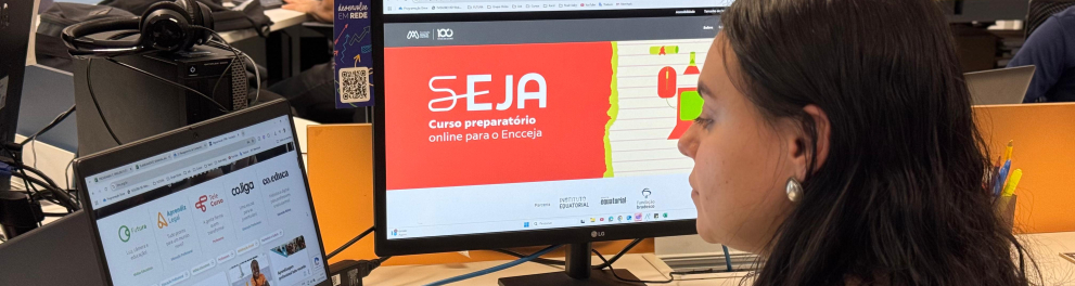 Uma mulher de cabelos longos e escuros, usando brinco de prata, está sentada em frente a duas telas em um ambiente de escritório. Ela olha para um monitor onde está aberto o site do curso “SEJA – Curso preparatório online para o Encceja”. À sua esquerda, há um notebook com a página inicial da Plataforma Futura aberta, exibindo programas como Co.liga, Telê Curso e Aprendê Legal. No fundo, outras pessoas também estão estudando. O ambiente é moderno, com iluminação clara e clima de concentração.