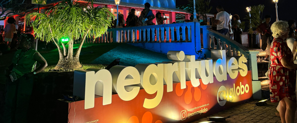 Foto mostra um letreiro iluminado em primeiro plano ("Negritudes"). Ao fundo, um jardim e, por fim, um casarão azul iluminado. É noite.