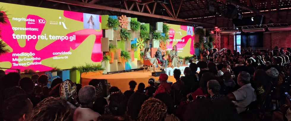 Auditório lotado assiste ao painel "Tempo Rei, Tempo Gil", durante o evento Negritudes Globo. No palco, decorado com plantas e painéis coloridos, estão sentados Gilberto Gil, a jornalista Maju Coutinho e a cantora Teresa Cristina, que conduzem a conversa. Um telão ao fundo exibe o nome do painel, os nomes dos participantes e as marcas dos organizadores. O ambiente é coberto, com estrutura de madeira e iluminação quente. 