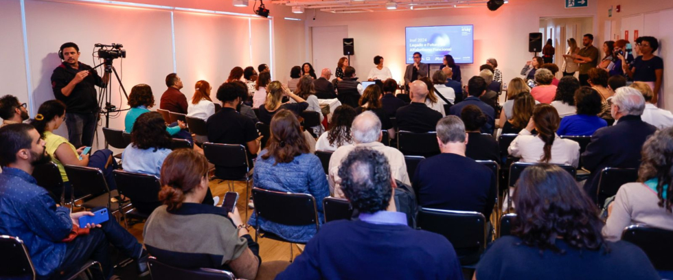 Uma sala cheia de pessoas assistindo uma apresentação. Tem pessoas sentadas de frente para o público. 