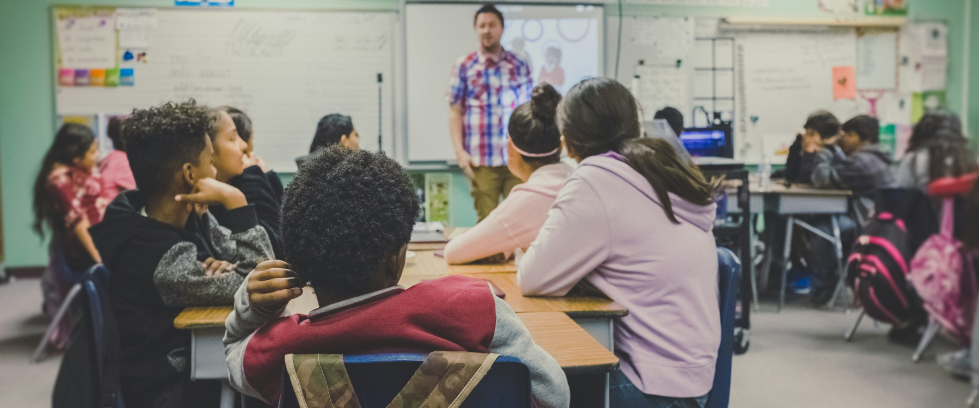 Sala de aula com diversos estudantes sentados em grupos ao redor de mesas, atentos ao professor que está em pé à frente da sala, próximo a um quadro branco com projetor. O ambiente é colorido e decorado com cartazes e materiais didáticos. Os alunos, de diferentes origens, estão concentrados na explicação, demonstrando engajamento com a atividade.