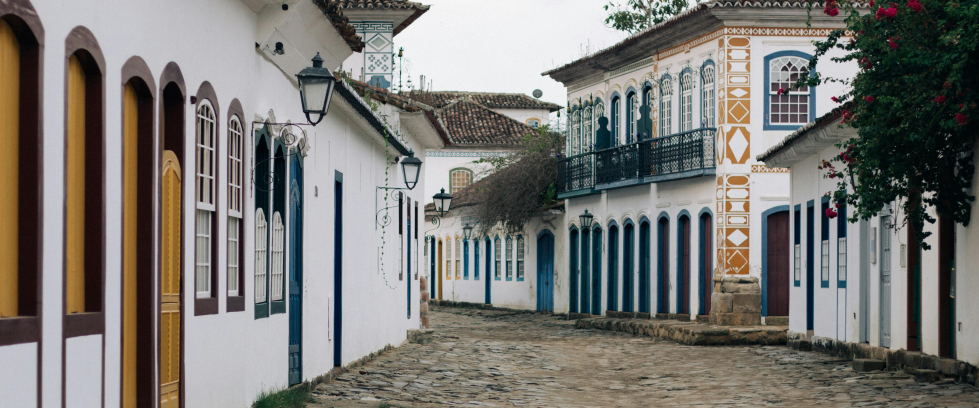Uma rua de pedras irregulares, típica do centro histórico de Paraty, no Rio de Janeiro, aparece vazia em um dia nublado. De ambos os lados, há casarões coloniais bem preservados, com paredes brancas, portas e janelas coloridas em tons como amarelo, azul, verde e vermelho. As janelas têm grades e caixilhos de madeira, e há postes de luz com design antigo fixados nas fachadas. Um dos prédios se destaca com detalhes ornamentais em amarelo e branco e sacada de ferro. À direita, uma trepadeira com flores vermelh