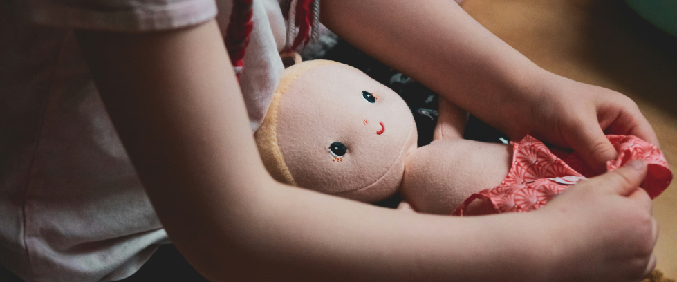 Criança branca sentada com boneca de pano no colo. O rosto da menina não é mostrado na imagem.