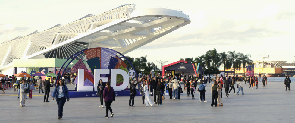 Vista ampla da área externa do Museu do Amanhã, no Rio de Janeiro, durante o Festival LED – Luz na Educação. Em primeiro plano, pessoas caminham e interagem ao redor de uma grande instalação colorida com as letras “LED”. Ao fundo, o mar, palmeiras e tendas coloridas compõem o cenário festivo.