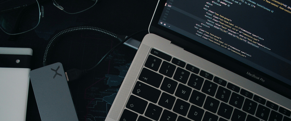 Um notebook visto parcialmente, aberto e exibindo uma tela com código de programação. O teclado preto com teclas iluminadas está em destaque. Ao lado do notebook, há um adaptador USB-C cinza conectado por um cabo. Também é possível ver um par de óculos, um caderno branco e um mapa-múndi estilizado como superfície da mesa. A iluminação é suave, sugerindo um ambiente de trabalho moderno e tecnológico.