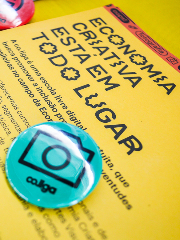 Material gráfico promocional sobre economia criativa disposto sobre uma superfície amarela. Em destaque, um folder amarelo com o título "ECONOMIA CRIATIVA ESTÁ EM TODO LUGAR" escrito com letras estilizadas. Abaixo, há um texto explicativo sobre a iniciativa Co.liga, uma escola livre digital gratuita voltada à inclusão produtiva de juventudes no campo da economia criativa. Em cima do folder, estão bottons coloridos, sendo o principal um azul com ícone de câmera e o nome "Co.liga", indicando uma abordagem vol
