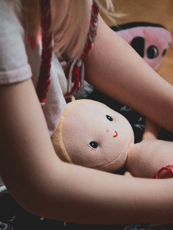 Criança branca sentada com boneca de pano no colo. O rosto da menina não é mostrado na imagem.