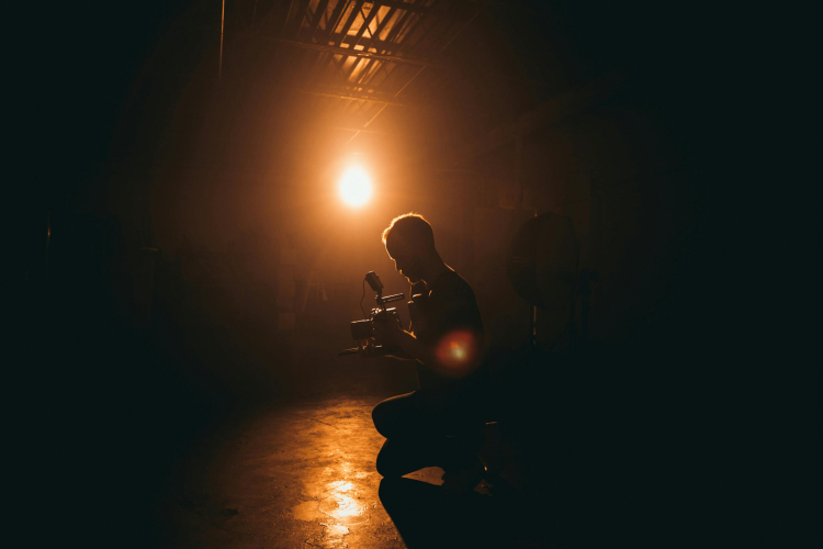 Em um ambiente escuro e ligeiramente esfumaçado (mais parece uma neblina, talvez), iluminado por uma lâmpada ao fundo, vê-se a silhueta de um homem agachadinho, segurando uma câmera.