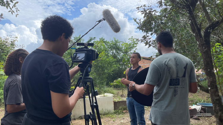 Equipe de filmagem trabalha em um ambiente com árvores e chão de terra.