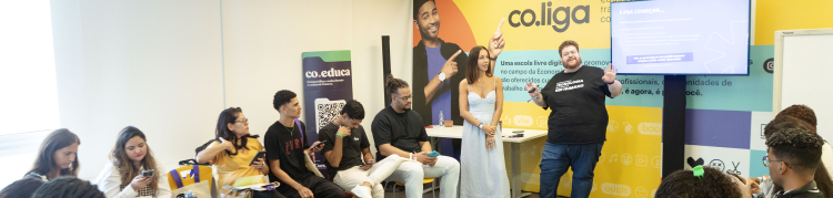 Cerca de 20 pessoas participam de uma oficina realizada em uma sala clara e colorida, com paredes decoradas com as marcas “co.liga” e “co.educa”. A maioria dos participantes está sentada em cadeiras amarelas e vermelhas, organizadas em semicírculo, com atenção voltada para dois facilitadores em pé ao centro — uma mulher de vestido claro e um homem com camiseta preta onde se lê “Transformando realidades”. Muitos usam celulares ou fazem anotações. À direita, uma tela exibe a frase “E pra começar...”, com inst